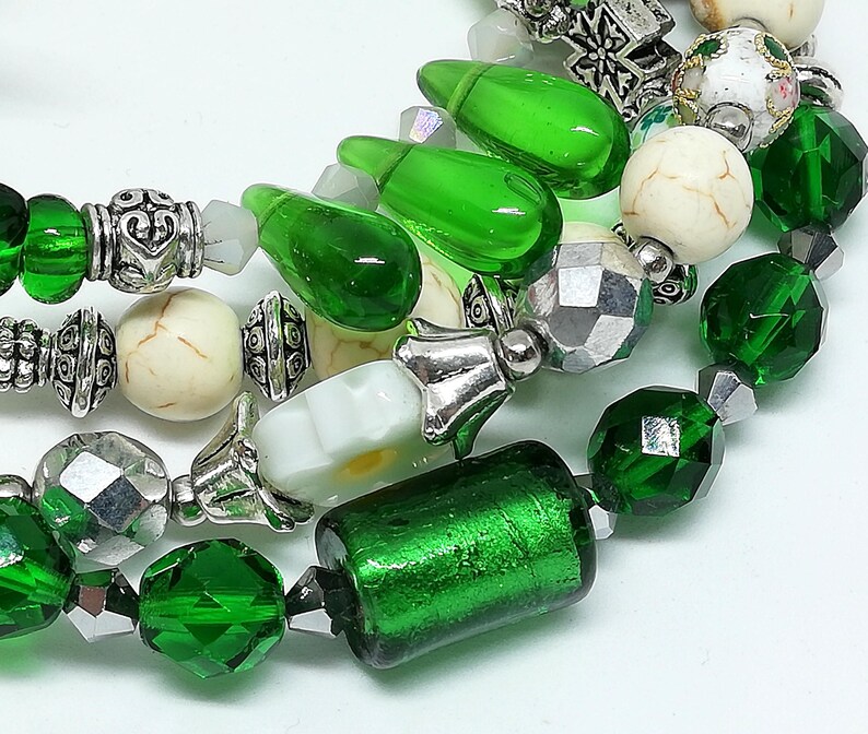 May include: Close-up of beaded jewelry featuring a mix of green, silver, and cream-colored beads. The beads include faceted green crystals, cylindrical green glass beads, and round cream-colored beads with a marbled effect. The jewelry is strung on a clear cord.