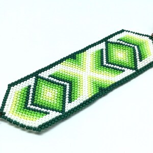 May include: Handmade beaded bracelet featuring a geometric design in shades of green, white, and yellow. The bracelet is rectangular with a dark green border and a decorative clasp. The beads are small and tightly woven.
