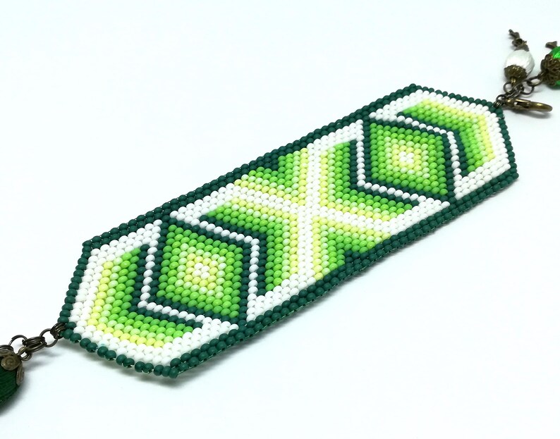 May include: Handmade beaded bracelet featuring a geometric design in shades of green, white, and yellow. The bracelet is rectangular with a dark green border and a decorative clasp. The beads are small and tightly woven.