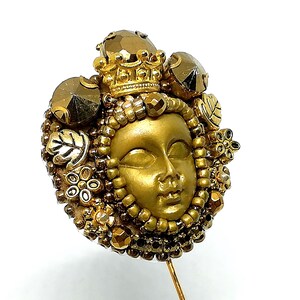 May include: A gold-toned decorative pin featuring a sculpted face with a crown and surrounding embellishments. The pin includes faceted gems, beads, and floral and leaf accents. The pin is set on a gold-colored metal pin.