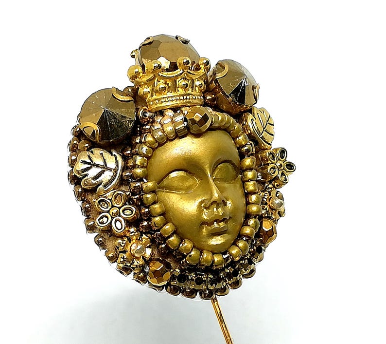 May include: A gold-toned decorative pin featuring a sculpted face with a crown and surrounding embellishments. The pin includes faceted gems, beads, and floral and leaf accents. The pin is set on a gold-colored metal pin.