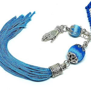 May include: A close-up of a blue tassel with silver-colored metal accents and decorative blue beads. The tassel is made of many strands of blue thread. A silver hand charm is also visible.