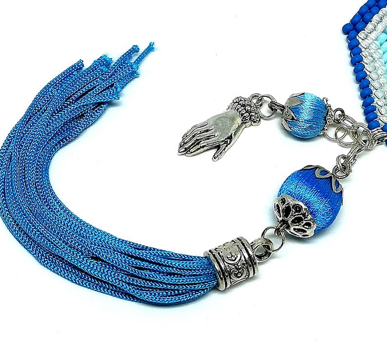 May include: A close-up of a blue tassel with silver-colored metal accents and decorative blue beads. The tassel is made of many strands of blue thread. A silver hand charm is also visible.