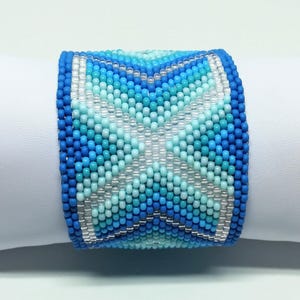 May include: A wide beaded bracelet in shades of blue and white. The bracelet features a geometric pattern with a central star-like design. The beads are small and tightly woven, creating a textured surface. The bracelet is displayed on a white surface.