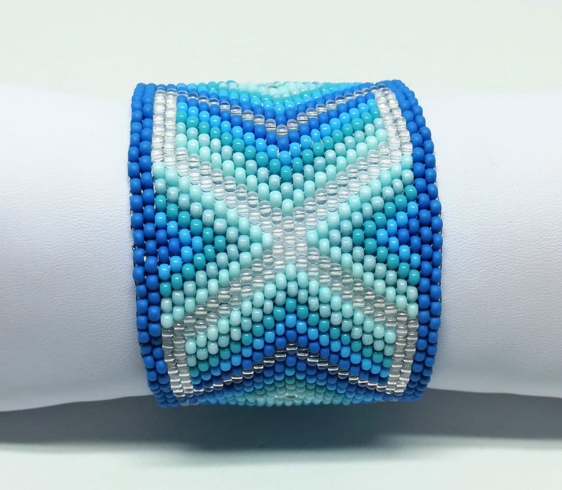 May include: A wide beaded bracelet in shades of blue and white. The bracelet features a geometric pattern with a central star-like design. The beads are small and tightly woven, creating a textured surface. The bracelet is displayed on a white surface.