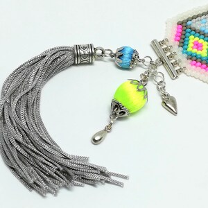 May include: A detailed close-up of a beaded bracelet with a silver tassel and decorative charms. The bracelet features a geometric pattern in pink, yellow, blue, and gray beads. The tassel is silver, and the charms include a heart and a neon green and yellow bead.