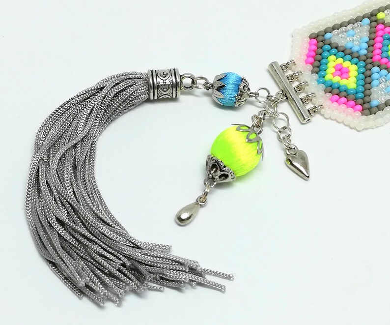 May include: A detailed close-up of a beaded bracelet with a silver tassel and decorative charms. The bracelet features a geometric pattern in pink, yellow, blue, and gray beads. The tassel is silver, and the charms include a heart and a neon green and yellow bead.