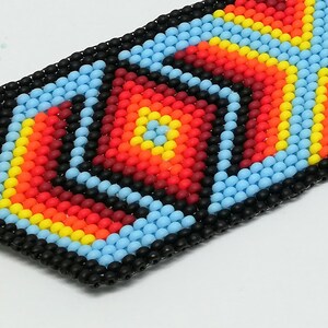 May include: Close-up of a beaded bracelet with a geometric design. The bracelet features a pattern of red, orange, yellow, and light blue beads, outlined in black. The design is centered with a square and surrounded by diamond shapes.