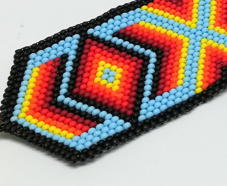 May include: Close-up of a beaded bracelet with a geometric design. The bracelet features a pattern of red, orange, yellow, and light blue beads, outlined in black. The design is centered with a square and surrounded by diamond shapes.