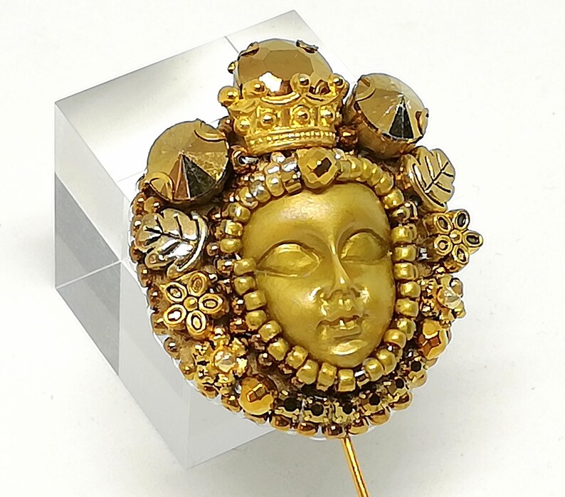 May include: A gold-toned brooch featuring a sculpted face with a crown and surrounding embellishments. The brooch is adorned with beads, floral accents, and leaf details. The design is set against a clear, rectangular base.
