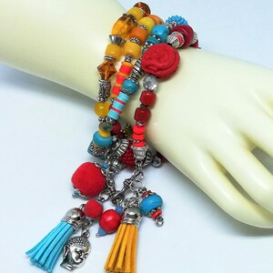 K&ouml;nnte beinhalten: Ein mehrreihiges Armband mit bunten Perlen, darunter Gelb, Orange, T&uuml;rkis und Rot. Das Armband hat eine rote geschnitzte Perle, silberne Charms und baumelnde Quasten in Blau und Gelb. Auf hellem Hintergrund.