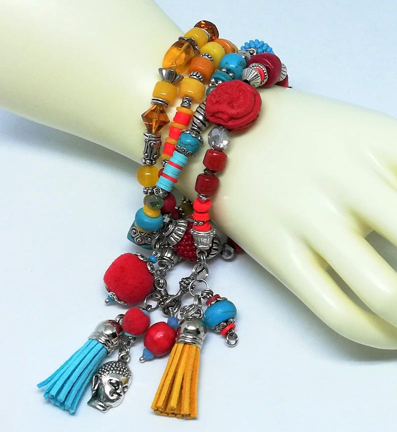 K&ouml;nnte beinhalten: Ein mehrreihiges Armband mit bunten Perlen, darunter Gelb, Orange, T&uuml;rkis und Rot. Das Armband hat eine rote geschnitzte Perle, silberne Charms und baumelnde Quasten in Blau und Gelb. Auf hellem Hintergrund.