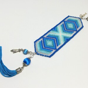 May include: A beaded bookmark with a geometric design in shades of blue and silver. The bookmark has a tassel on one end and decorative charms on the other. The design features an X and diamond pattern.