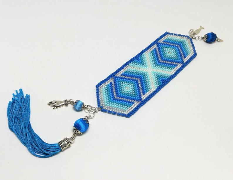 May include: A beaded bookmark with a geometric design in shades of blue and silver. The bookmark has a tassel on one end and decorative charms on the other. The design features an X and diamond pattern.