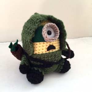 May include: A crocheted minion toy wearing a green hooded cloak and holding a brown and green bow and arrow. The minion has a yellow body, a green eye patch, and a black mouth.