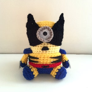 Op de afbeelding: Een geel gehaakt minion-speelgoed met een zwart masker en een rode riem. De minion heeft één groot grijs oog en zit op een wit oppervlak.