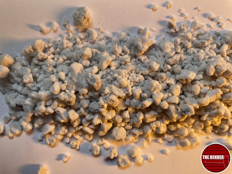 May include: A collection of small, irregular, off-white, porous, and textured mineral-like granules scattered across a surface. The pieces vary in size and shape, exhibiting a rough, uneven surface structure. A circular logo with the text THE BENDER is visible.