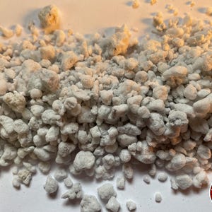 May include: A pile of white perlite granules with irregular shapes and porous textures. The small, lightweight volcanic glass particles are scattered across the frame. A circular red logo with white horizontal lines and the text THE BENDER is visible.