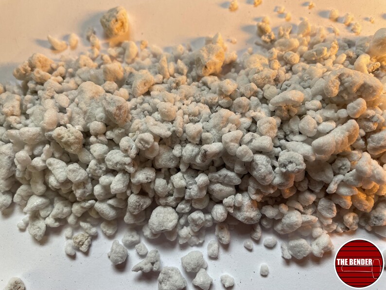 May include: A pile of white perlite granules with irregular shapes and porous textures. The small, lightweight volcanic glass particles are scattered across the frame. A circular red logo with white horizontal lines and the text THE BENDER is visible.