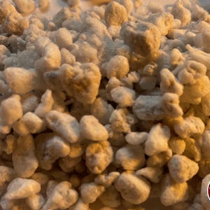 May include: A collection of small, irregular, off-white and light beige mineral granules with a porous, textured surface. The particles vary in size and shape, creating a dense, uneven pile. A circular logo with the text THE BENDER is visible.