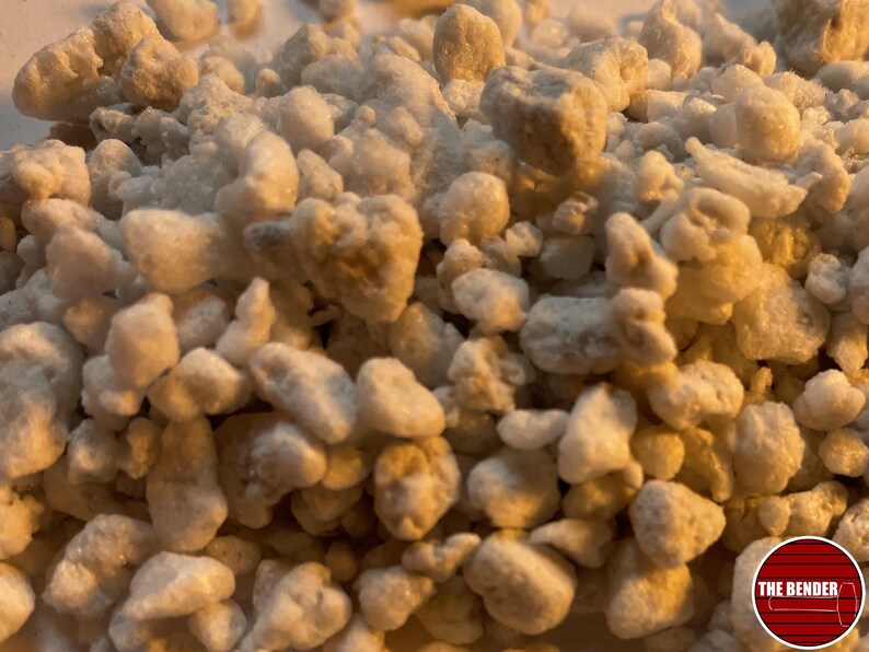 May include: A collection of small, irregular, off-white and light beige mineral granules with a porous, textured surface. The particles vary in size and shape, creating a dense, uneven pile. A circular logo with the text THE BENDER is visible.