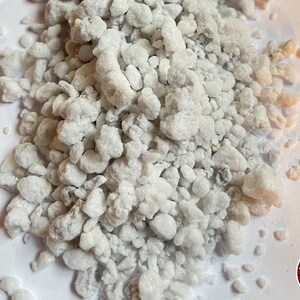 May include: A pile of white, porous, irregular perlite granules with a rough, rocky texture. In the bottom right corner, there is a circular red logo containing horizontal black lines and the white text THE BENDER.
