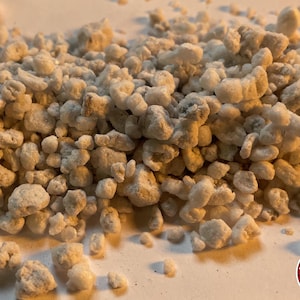 May include: A collection of small, irregular, off-white and light gray porous mineral granules. The texture is rough and uneven, with varying sizes of individual particles scattered across the frame. A circular logo with the text THE BENDER is visible.