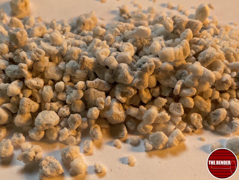 May include: A collection of small, irregular, off-white and light gray porous mineral granules. The texture is rough and uneven, with varying sizes of individual particles scattered across the frame. A circular logo with the text THE BENDER is visible.