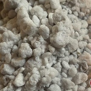 May include: A collection of small, irregular white and off-white mineral stones with a crystalline, textured surface. The stones vary in size and shape, creating a dense, granular pile. A circular red logo with the text THE BENDER is visible.