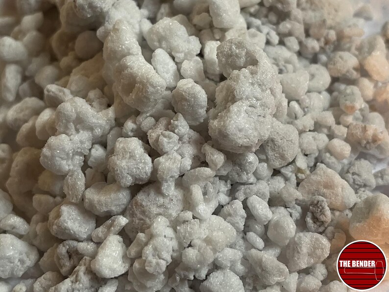 May include: A collection of small, irregular white and off-white mineral stones with a crystalline, textured surface. The stones vary in size and shape, creating a dense, granular pile. A circular red logo with the text THE BENDER is visible.
