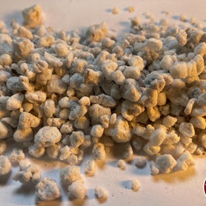 May include: A collection of small, irregular, off-white perlite granules with a porous, lightweight texture. The particles vary in size and shape, displaying a matte finish. A circular logo in the bottom right corner features the text THE BENDER.