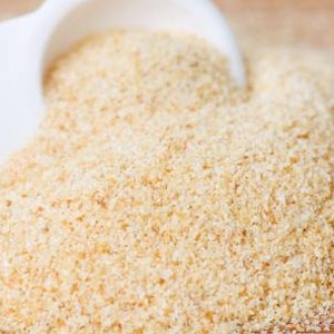 May include: A white spoon filled with granulated garlic powder, spilling onto a wooden surface.