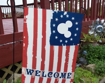 May include: A welcome garden flag with a distressed American flag design. The flag features a white Mickey Mouse silhouette in the blue field of stars. The words "WELCOME" are printed in white on the red stripes.