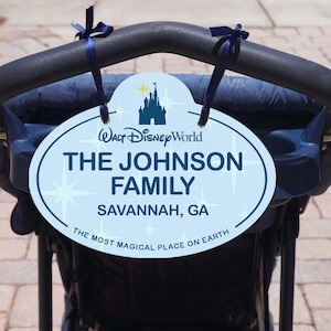 May include: A light blue oval sign with the text "Walt Disney World," "The Johnson Family," and "Savannah, GA." The sign is attached to a stroller handle with blue ribbons. The bottom of the sign reads "The Most Magical Place on Earth."