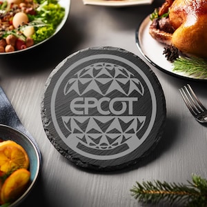 May include: A black slate coaster with a gray engraved design of the Epcot logo, featuring the iconic Spaceship Earth sphere.