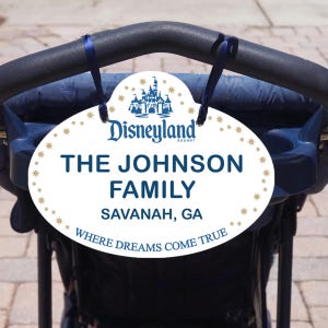 May include: An oval white Disneyland Resort sign with the text "THE JOHNSON FAMILY, SAVANAH, GA" in navy blue. The sign is decorated with gold stars and a navy blue castle graphic. The text "WHERE DREAMS COME TRUE" is at the bottom.