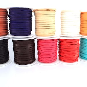 Op de afbeelding: Tien spoelen met lederen koord in verschillende kleuren, waaronder oranje, paars, beige, wit, bruin, zwart, roze, rood, turquoise en bruin. De spoelen zijn in een rij op een wit oppervlak gerangschikt.