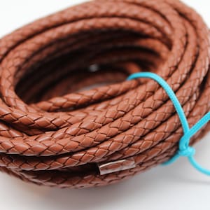 May include: A close-up of a brown braided leather cord, tied with a turquoise string. The cord is coiled in a circle and is approximately 5/16 inch thick.