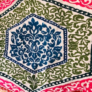 May include: Close-up of a textile with an intricate embroidered design. The pattern features a central blue floral motif within a white hexagon, surrounded by green and pink geometric shapes. The embroidery is detailed and colourful.