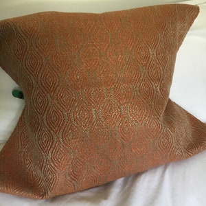 May include: Square decorative pillow with an orange and gold diamond pattern. The pillow has a textured appearance and is resting on a white surface. The pillow is a home decor item.