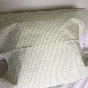 Könnte beinhalten: Ein hellgrüner Kissenbezug mit einem sich wiederholenden Muster aus weißen Ovalen. Der Kissenbezug liegt auf einem weißen Bett, wobei ein Teil umgeschlagen ist. Der Stoff wirkt weich und das Design ist schlicht.