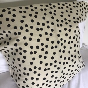 May include: A beige fabric with a repeating pattern of black polka dots. The fabric is draped over a white surface, with the edges of the fabric visible. The polka dots are evenly spaced and vary slightly in size.