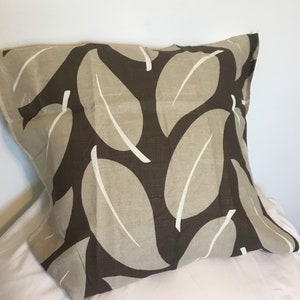 May include: Square decorative pillow with a leaf pattern. The pillow features large, stylized leaves in shades of brown and beige, with white accents. The pillow is resting on a white surface.