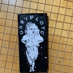 Puede incluir: Imagen en blanco y negro de una persona con una peluca afro grande y un vestido blanco. El texto "GARY FLOYD" está escrito en un círculo alrededor de la persona.