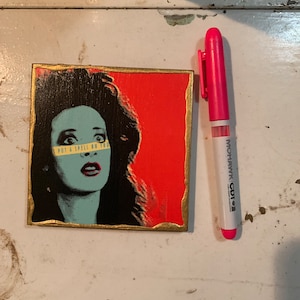 Op de afbeelding: Een vierkant, ingelijst schilderij met een gouden rand. Het schilderij toont een vrouw met donker haar en een blauwgroen gezicht. De vrouw draagt een geel en zwart gestreept band over haar ogen. De tekst "Put a Spell on You" staat op het band. De achtergrond van het schilderij is rood.