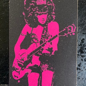 Op de afbeelding: Een roze silhouet van een persoon die een elektrische gitaar bespeelt. De persoon heeft groot, krullend haar en draagt een kort rokje en een top met een decoratief ontwerp. De gitaar is een elektrische gitaar met een massief lichaam en een enkele cutaway.