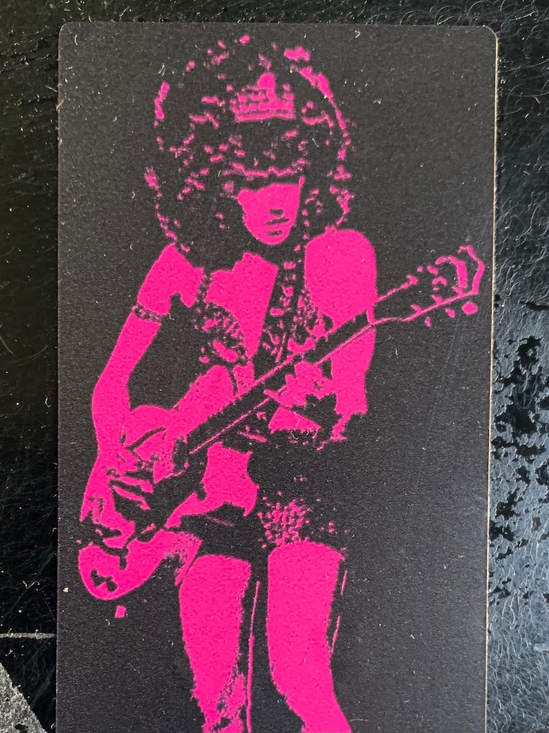 Op de afbeelding: Een roze silhouet van een persoon die een elektrische gitaar bespeelt. De persoon heeft groot, krullend haar en draagt een kort rokje en een top met een decoratief ontwerp. De gitaar is een elektrische gitaar met een massief lichaam en een enkele cutaway.