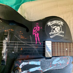 Op de afbeelding: Een zwarte elektrische gitaar met verschillende stickers, waaronder een roze sticker van een vrouw die gitaar speelt, een zwart-witte sticker van een schedel en gekruiste beenderen met een kat met een ooglapje en een blauw-witte sticker van een vrouw in een rok.