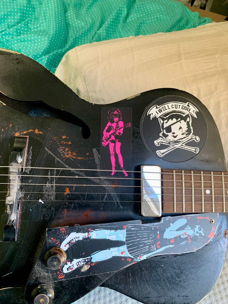 Op de afbeelding: Een zwarte elektrische gitaar met verschillende stickers, waaronder een roze sticker van een vrouw die gitaar speelt, een zwart-witte sticker van een schedel en gekruiste beenderen met een kat met een ooglapje en een blauw-witte sticker van een vrouw in een rok.