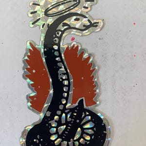 May include: A holographic sticker featuring a black snake with a white halo above its head and orange wings on its sides. The snake is coiled at the bottom and has a detailed scale pattern. The entire design has a reflective, prismatic finish.